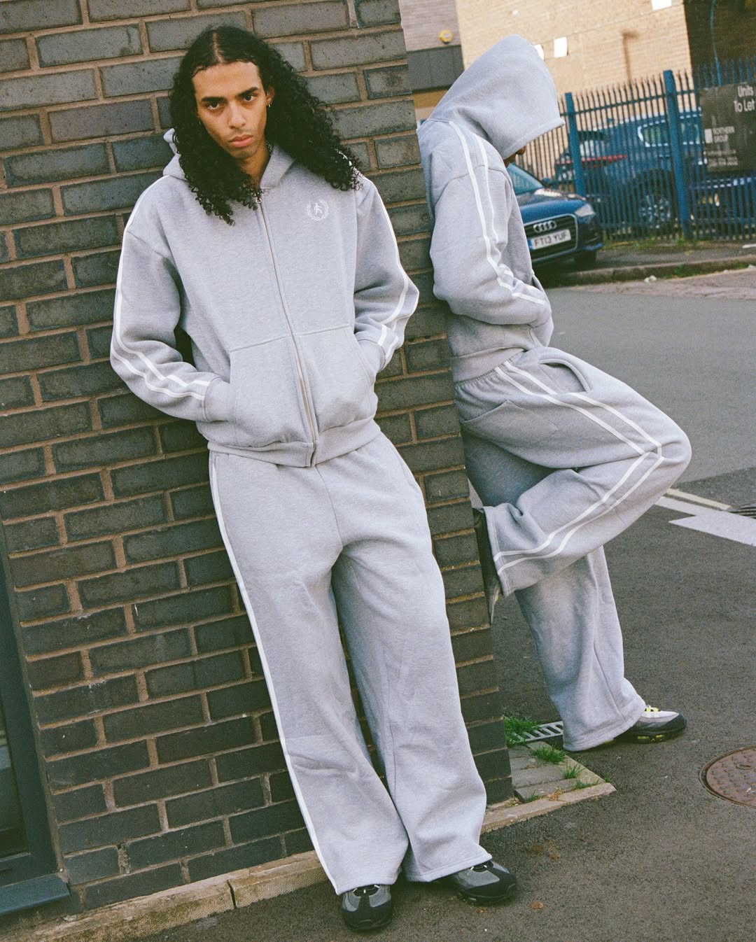 Two models wearing the Grey Sportacus hoodie and joggers from Ewubawn’s Akimbo collection, leaning casually against a brick wall.